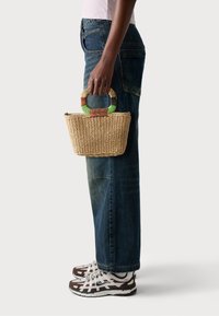 Woven straw basket bag with a multicolored handle, rectangular shape, and natural texture, being held beside denim jeans and patterned sneakers.