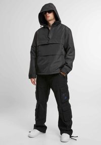 Jachetă cu glugă neagră, tip pulover, cu fermoar pe jumătate și buzunar frontal tip plic, purtată împreună cu pantaloni cargo cu multiple buzunare și adidași albi.
