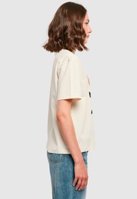 Cream-colored cotton t-shirt with short sleeves and a relaxed fit, featuring a bold black graphic on the front. Paired with light blue jeans.