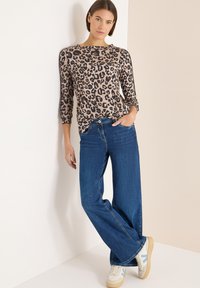 Bluzka w panterkę z rękawami 3/4, zestawiona z szerokimi, niebieskimi jeansami o szerokich nogawkach. Modelka ma na sobie beżowo-niebieskie sneakersy. Gładka tekstura.