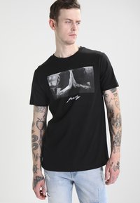 Camiseta de algodón de manga corta en color negro, con un gráfico frontal de manos entrelazadas y el texto "reza". Corte casual con un escote redondo.