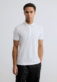 Λευκό πόλο μπλουζάκι από βαμβάκι, με πλάκα δύο κουμπιών, κοντά μανίκια και ανοιχτό μπλε λεπτομέρειες στον γιακά και στα μανίκια.