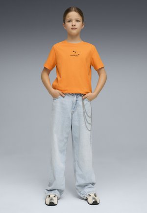 Bambino in piedi con le mani nelle tasche dei jeans azzurri, indossando una maglietta arancione Puma McLaren e sneakers bianche su sfondo grigio.