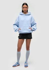Felpa oversize azzurro chiaro con tasca frontale, mini gonna cargo nera, calze fino al ginocchio e sneaker blu con dettagli bianchi.