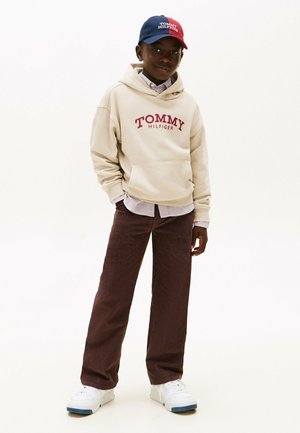 Beige hoodie met het "TOMMY HILFIGER" logo, gelaagd over een gestreept shirt. Bruine corduroy broek en witte sneakers maken de outfit compleet. Blauwe en rode pet.