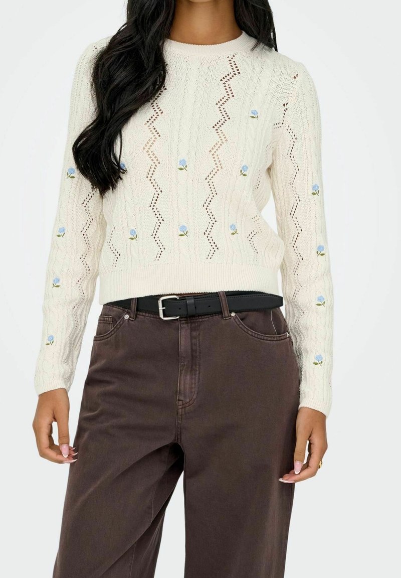 Femme portant un pull en maille crème avec de petites broderies de fleurs bleues, associé à un pantalon marron foncé taille haute et une ceinture noire.