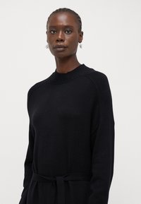 Fekete kötött ruha bordázott nyakkivágással, hosszú ujjal és derékon megkötős övvel. Sima textúra, minimalista dizájn.