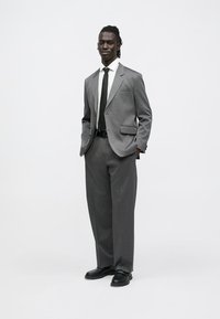 Hall hall ülikond mustriga tekstuuriga, laiad püksid, sobiv lips ja mustad kingad. Komponendid hõlmavad kahe nupuga bleiserit ja valget särki.