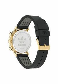 Boîtier de montre en acier inoxydable doré avec un bracelet en caoutchouc noir perforé. L'arrière affiche le logo Adidas et les détails du mouvement.