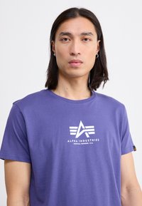 Purpurinė medvilninė marškinėliai su trimis dryžiais ir tekstu "ALPHA INDUSTRIES" centrine dalimi.
