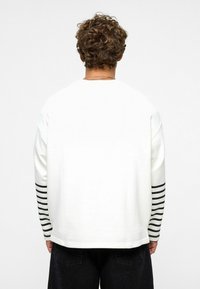 Witte langemouwen shirt met zwarte horizontale strepen op de mouwen. Zachte stof, relaxte pasvorm, ronde hals en minimalistisch design.