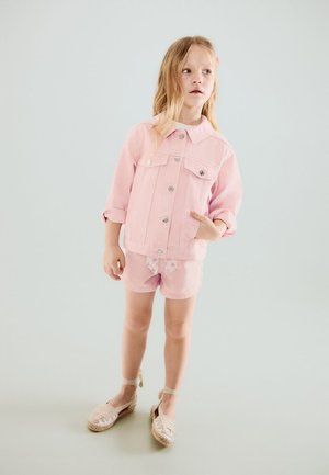 Jong meisje met lang blond haar, een roze jas, roze short met een strikje met sterrenpatroon en beige espadrilles.