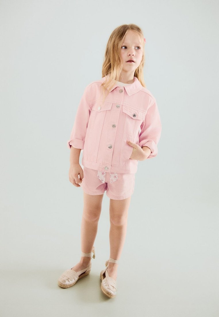 Jeune fille aux longs cheveux blonds portant une veste rose, un short rose avec un nœud à motif d'étoiles et des espadrilles beiges.