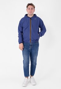 Giacca leggera blu con cappuccio e zip gialla, due tasche laterali e polsini elastici, abbinata a jeans blu e sneakers bianche.