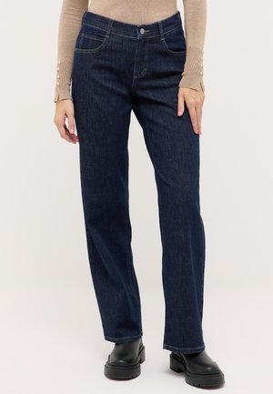Person iført mørkeblå jeans med lige ben, sorte støvler og en beige langærmet top med knappedetaljer, stående mod en lys baggrund.