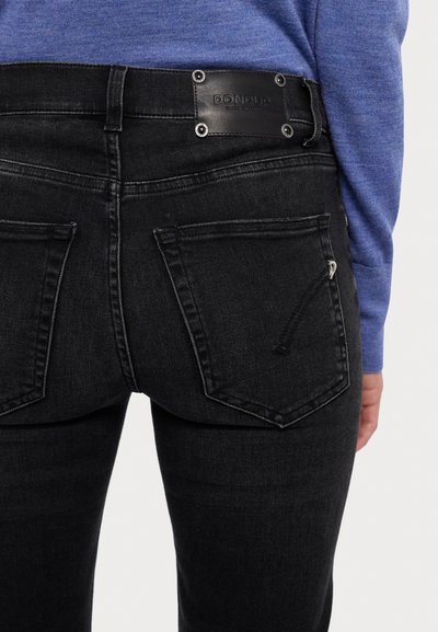 Jean en denim noir avec une étiquette en cuir portant un texte en relief, deux poches arrière et une coupe slim. Le tissu a un léger éclat.