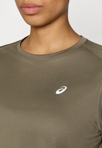 Αθλητική μπλούζα ελιάς με στρογγυλή λαιμόκοψη, λεία υφή και αντανακλαστικό λευκό λογότυπο στο αριστερό στήθος.