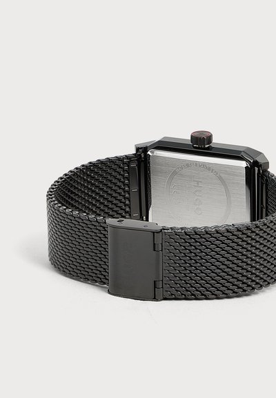 Bracelet de montre en maille métallique noire avec plaque arrière argentée rectangulaire et fermoir noir, vu de dos sur fond clair.