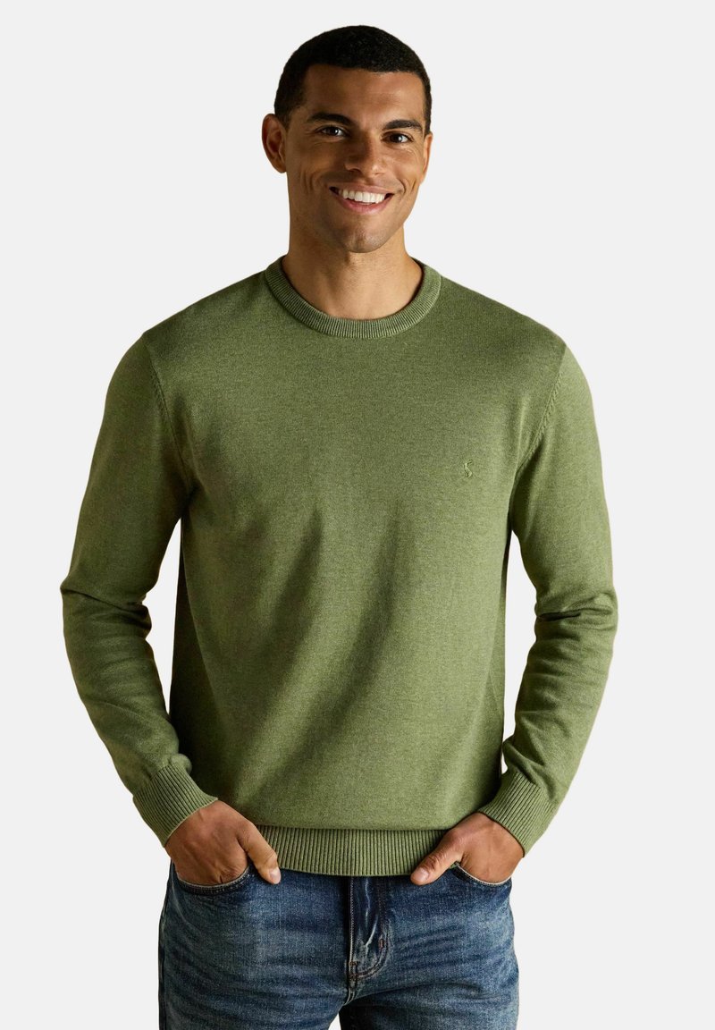 Muž sa usmieva, nosí olivovo zelený sveter s okrúhlym výstrihom a modré rifle, ruky čiastočne vreckách, na svetlom jednoduchom pozadí.