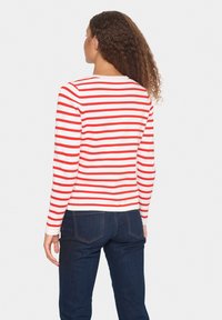 Chemise à manches longues, blanche avec des rayures horizontales rouges, dotée d'un col rond. Portée avec un jean en denim foncé. Matière en coton lisse.