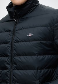 Personne portant une veste matelassée noire avec un col montant et une fermeture éclair à l'avant, avec un petit logo GANT brodé sur la poitrine.