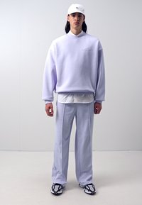 Gaiši purpursarkans pārpasaulīgs krekls ar izšuvumu, pārlikts pār baltu krekli, saskanīgi savienots ar gaiši purpursarkanu biksēm un zīmētiem sporta apaviem.