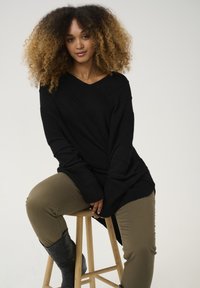 Maglione oversize nero con texture, scollo a V e maniche lunghe, abbinato a pantaloni aderenti verde oliva e stivaletti in pelle nera.