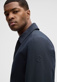 Jonge man in een donkerblauwe jas met een subtiel geperst logo op de bovenste mouw, kijkt over zijn schouder tegen een effen lichte achtergrond.