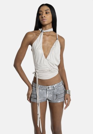 Kvinde iført hvid halterneck-top med dyb V-udskæring og bindebånd i taljen, kombineret med sølvfarvede metallic shorts og et sølvarmbånd på hendes højre håndled.