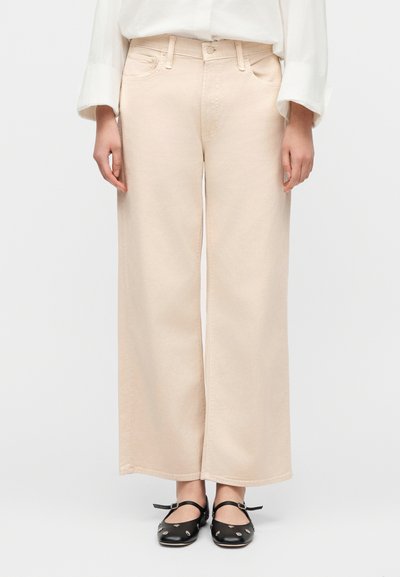 Άτομο που φοράει μπεζ παντελόνι με φαρδιά μπατζάκια και μαύρα παπούτσια Mary Jane με μεταλλικά τρουκς, στέκεται μπροστά σε απλό λευκό φόντο.