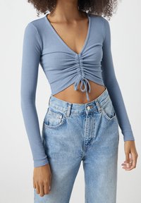 Blau gestreifter Langarm-Crop-Top mit tiefem V-Ausschnitt und gerafftem Frontbereich. Kombiniert mit hoch taillierten hellblauen Denim-Jeans.