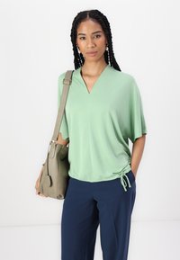 Blusa a maniche corte, con scollo a V, in tessuto verde chiaro con dettaglio a fiocco all'orlo. Abbinata a pantaloni blu navy e una borsa verde oliva.
