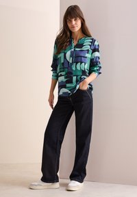Flerfärgad blus med geometriska former i grönt och blått, avslappnad passform, långa ärmar och knäppning, i kombination med mörka jeans och sneakers.