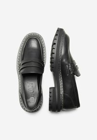 Mocasines de cuero negro con punta redondeada, acento de doble correa, suela de goma gruesa y detalle de costura blanca en contraste a lo largo de los bordes.