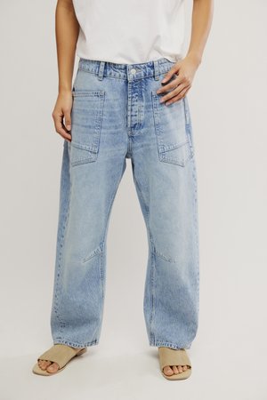 Person iført lyseblå vidde jeans med store forlommer, hvid top og beige åbne sandaler, der står foran en ensfarvet baggrund.