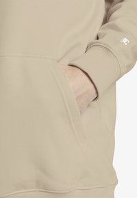 Felpa beige realizzata in morbido tessuto, presenta una tasca a marsupio e polsini a costine. Dettaglio del logo discreto sulla manica. Texture complessiva liscia.