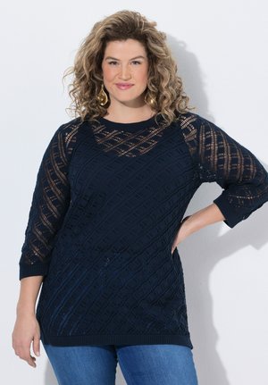 Krulligharige vrouw met gouden oorbellen draagt een marineblauwe textuurgebreide top en blauwe jeans, staand met één hand op haar heup.