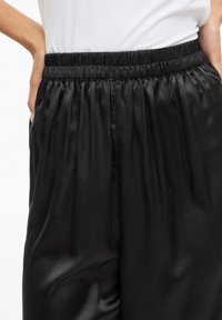 Czarne satynowe spodnie z elastycznym pasem, charakteryzujące się gładką fakturą i luźnym krojem, z zakładkami podkreślającymi design.