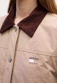 Veste beige avec col en velours côtelé marron, boutons argentés et écusson avec le logo "Tommy Jeans" sur la poitrine.