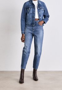 Jeansjacka med rhinestones, vit kort topp, blå högmidjade jeans och bruna klackiga ankelskor, står mot en neutral bakgrund.