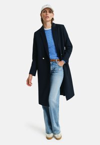 Manteau long navy avec deux boutons dorés, pull en tricot à torsades bleu clair, jeans évasés en denim clair et baskets blanches avec une semelle beige.
