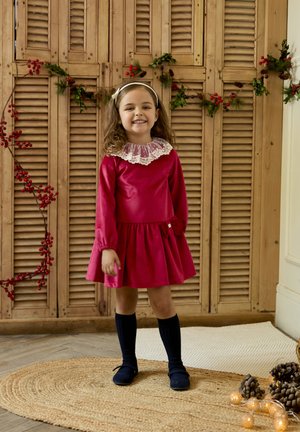 Vestido rojo de manga larga con un cuello de encaje y falda recogida, adornado con un lazo. Combinado con calcetas de rodilla azul marino y zapatos oscuros.