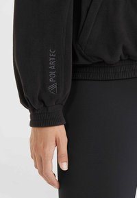 Pull-over en polaire noir avec des poignets élastiques, comportant un logo "Polartec" discret sur la manche, texture lisse et coupe décontractée.