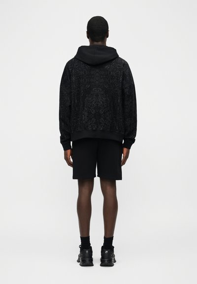 Bărbat purtând hanorac negru cu model, pantaloni scurți negri, șosete negre și adidași negri, stând cu fața spre spate pe un fundal simplu.