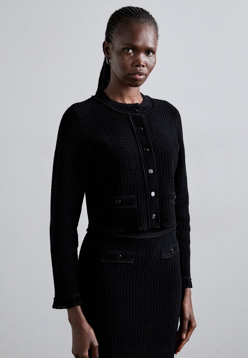 Mustakuvioinen neulehuppari, jossa on pyöreä kaula-aukko, pitkät hihnat ja etunapin sulkeminen. Sisältää läppätaskut napin yksityiskohdilla.