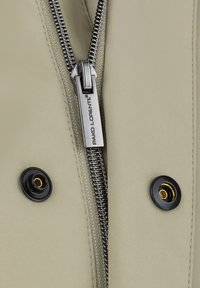 Jasnobeżowa kurtka z metalowym zamkiem błyskawicznym i czarnymi zatrzaskami. Materiał ma gładką teksturę i matowe wykończenie.