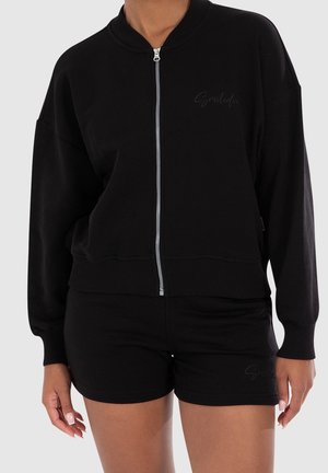 Vrouw draagt een zwarte bomberjas met ritssluiting en bijpassende zwarte korte broek, met subtiele cursieve tekst op de borst en de korte broek.