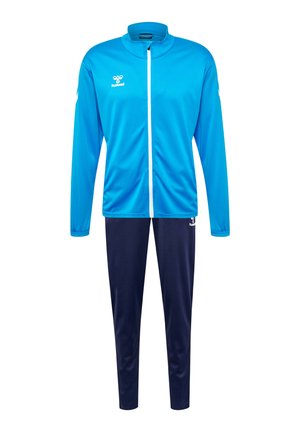 Blauwe sportjack met een hoge kraag en volledige rits, gecombineerd met marineblauwe broek. Glad materiaal, met logo op de borst en het broekbeen.