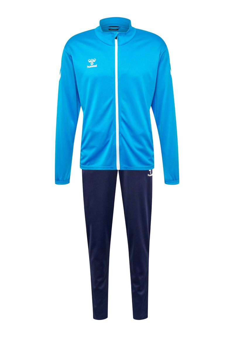Blaue Sportjacke mit hohem Kragen und durchgehendem Reißverschluss, kombiniert mit navyblauen Hosen. Glattes Material, mit Logo auf der Brust und am Hosenbein.