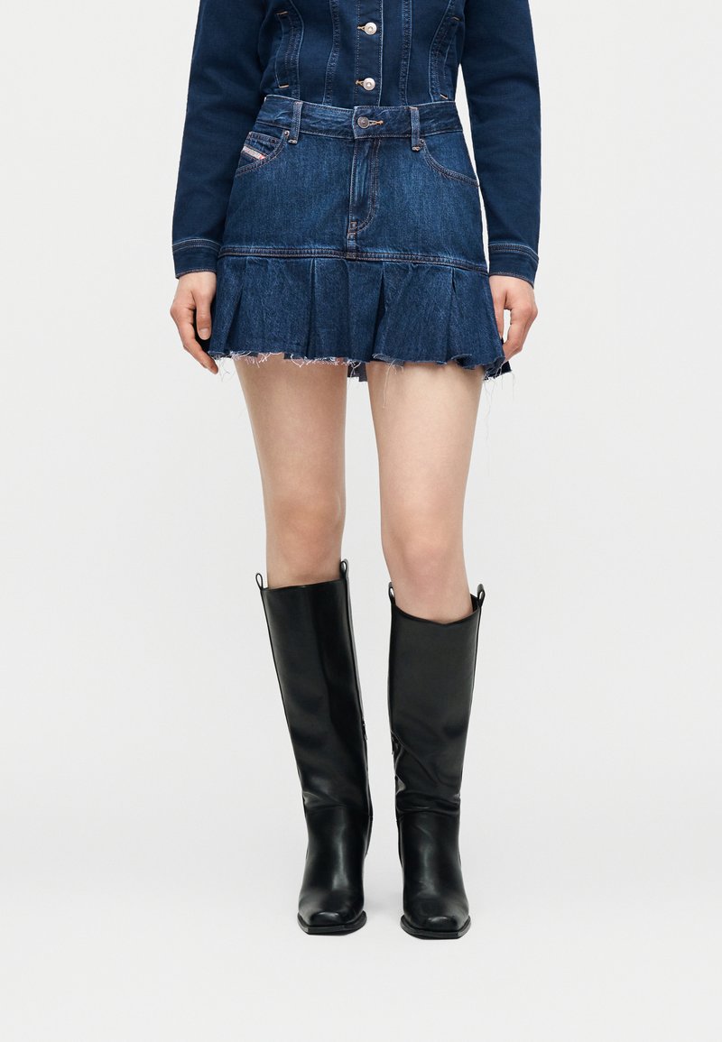 Άτομο που φοράει φούστα από τζιν με βολάν, τζιν μπουφάν που ταιριάζει και μαύρες δερμάτινες μπότες μέχρι το γόνατο μπροστά σε απλό φόντο.